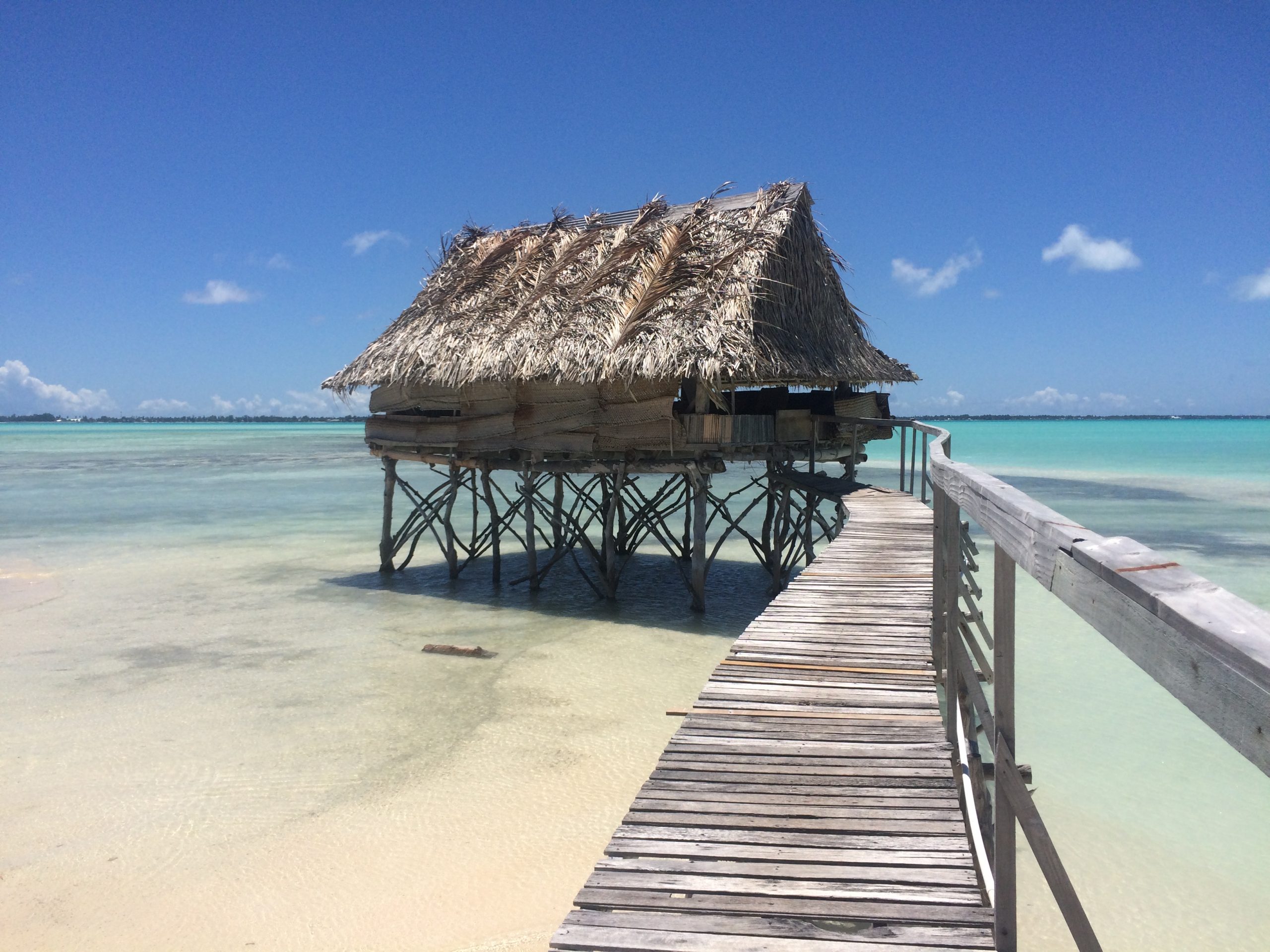 A picturesque view of a traditional Kiribati overwater buia, a thatched-roof hut built on stilts above the clear, turquoise waters of the Pacific Ocean. The buia is intricately constructed with local materials, showcasing Kiribati's traditional architecture. The image captures the unique charm of Kiribati's coastal living, highlighting the vibrant culture and connection to the surrounding natural beauty.