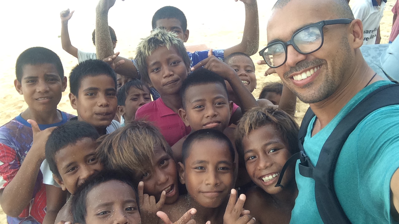 A heartwarming selfie featuring the user and cheerful Kiribati children on a sunny beach. The user is smiling, arm around the kids, capturing the genuine joy of the moment. The background shows the golden sands, sparkling ocean waves, and a clear blue sky, emphasizing the happiness shared between the me and the local children in Tarawa.