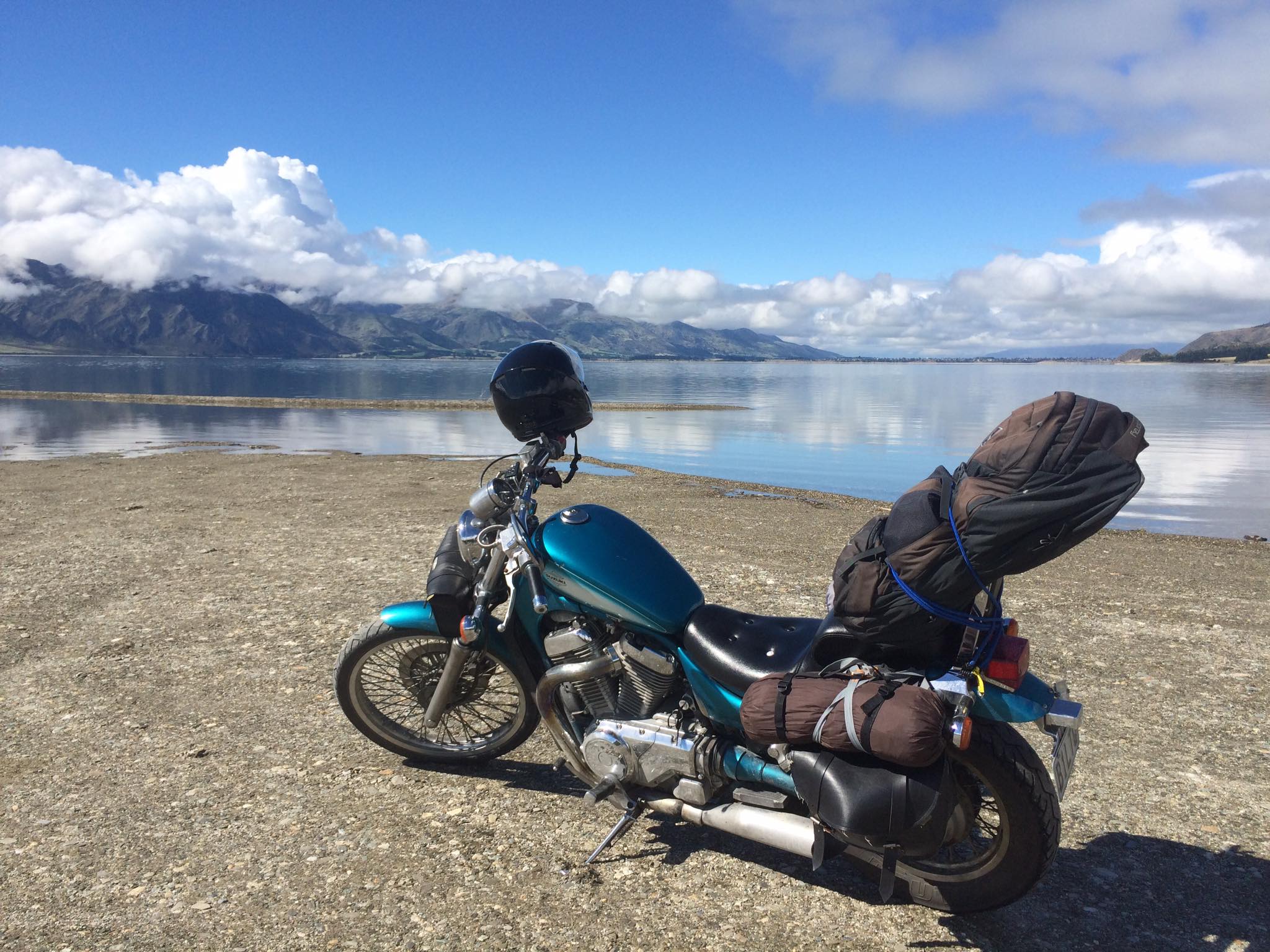 A captivating photo featuring a motorcycle parked near the tranquil shores of a lake in the South Island of New Zealand. The motorcycle, with its sleek design, contrasts against the serene waters of the lake, reflecting the surrounding mountains and the clear blue sky. The image captures the sense of adventure and freedom, illustrating a moment of peaceful contemplation amidst the breathtaking natural beauty of the South Island.