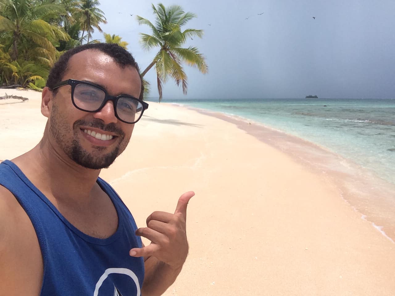 sandy beach, throwing a Shaka hand sign with a wide smile. In the background, the turquoise waters of the Pacific Ocean meet the clear blue sky, creating a serene tropical scene.