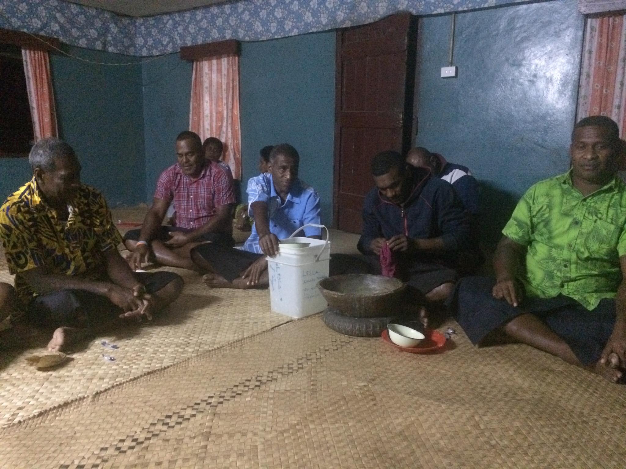 A vibrant scene of community togetherness in a Fijian village during a Kava night. Sitting in a circle, holding coconut shell cups filled with Kava, a traditional Fijian drink. The atmosphere is warm and convivial, reflecting the spirit of shared culture and camaraderie in Fiji